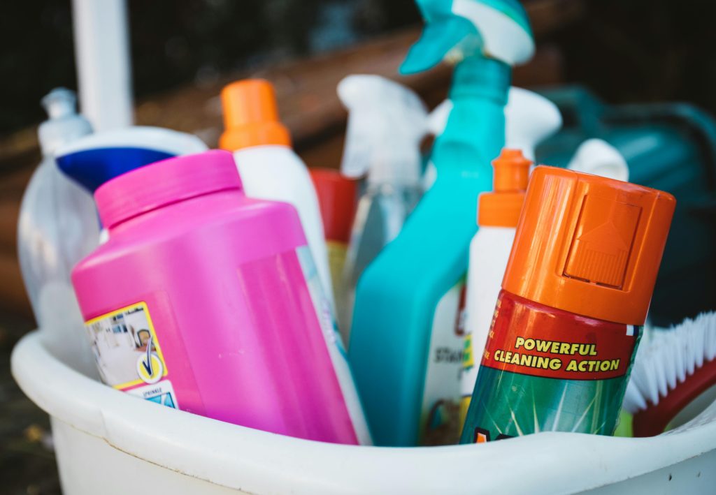 pexels-photo-3177257-3177257 Close-up of assorted cleaning supplies in a bucket outdoors, showcasing various detergents and sprays.